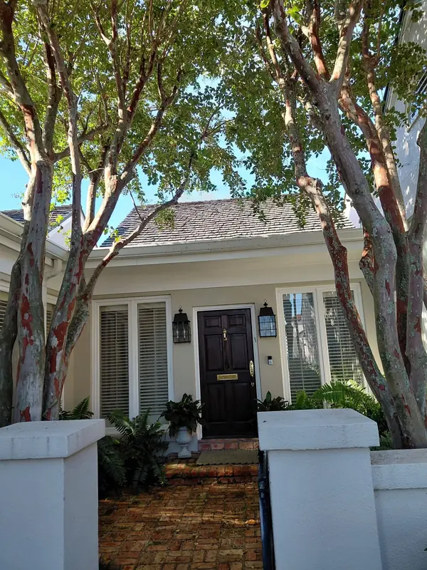 Pale yellow cottage with white shutters and a black front door between crape myrtle trees