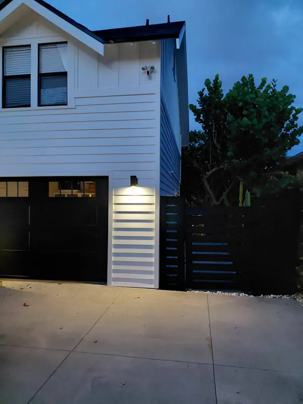 White board-and-batten house with a black garage door and slatted fence at dusk